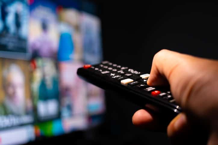 A hand holding a TV remote with the TV blurred out in the background.