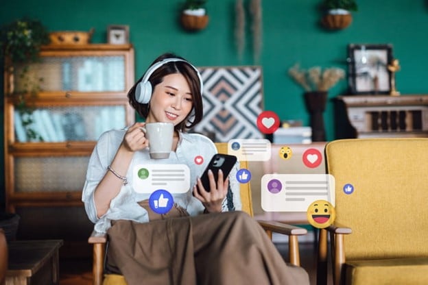 A person using a smartphone with social media notifications and emojis, holding a coffee mug while wearing headphones in a cozy room.