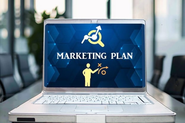 A laptop displaying "Marketing Plan" with icons of a person, magnifying glass, and graph in an office setting.