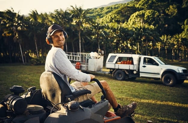 A worker on a ride-on mower with a truck and equipment in the background on a grassy area.