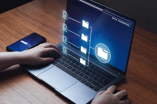 A person using a laptop with a holographic display showing "DATA TRANSFER..." and a sequence of file folders being moved, with a smartphone nearby on a wooden desk.