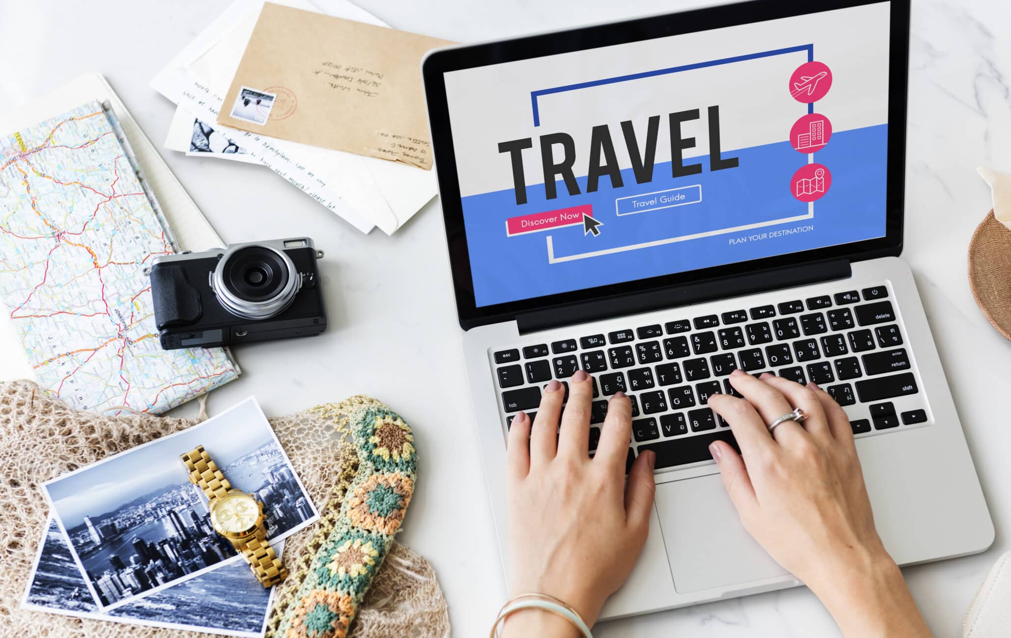 Laptop displaying a travel website during online trip planning, with maps, photos, and travel items arranged on a desk.