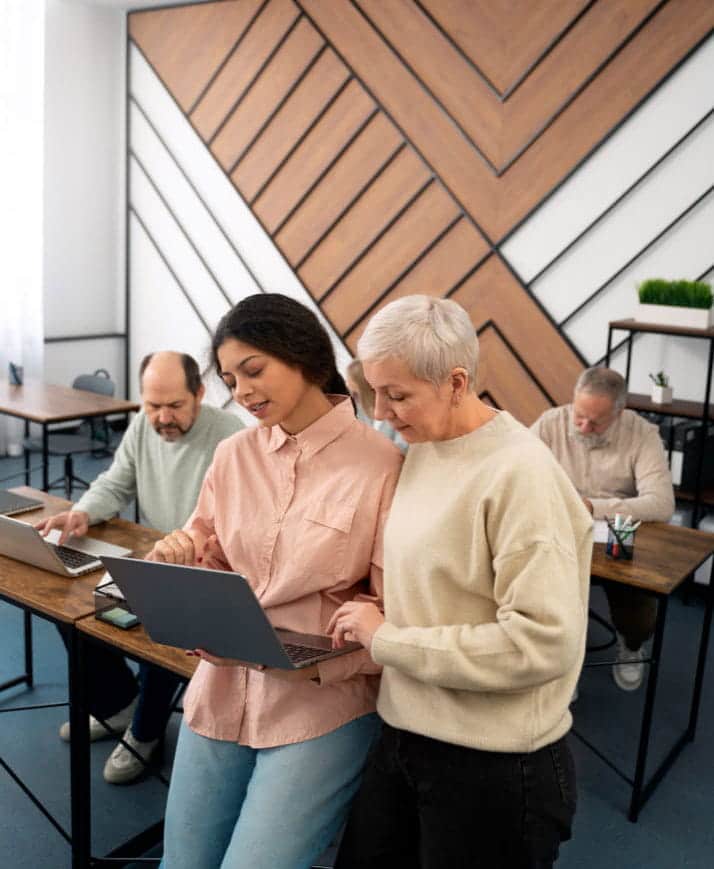 Senior adult learning to use a laptop with guidance in a modern classroom setting.